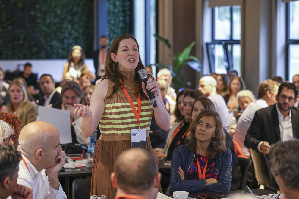 A room of people, looking at a person standing talking into a microphone